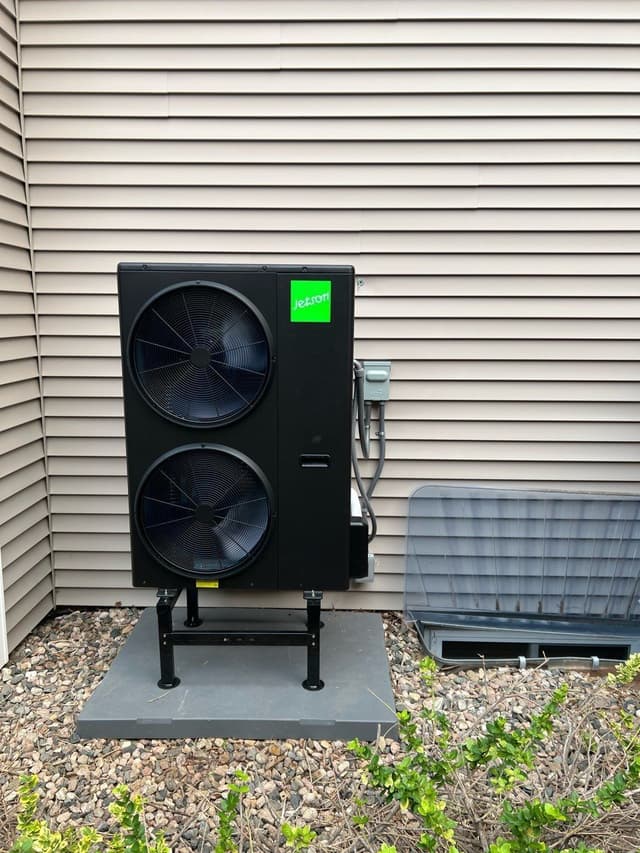 Jetson Air central ducted heat pump mounted on a metal stand over a concrete pad in a gravel bed along the side of a gray lap siding house in Fort Collins, CO, with electrical disconnect and conduit runs visible to the right.