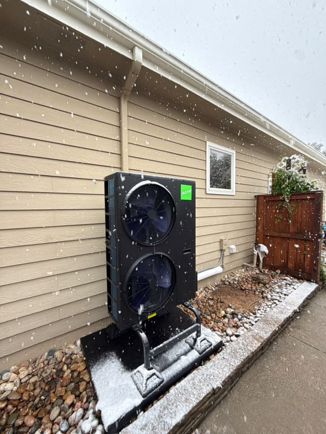 A black Jetson Air 4-ton heat pump on a raised stand in a gravel bed along the side of a tan house in Fort Collins, CO, with snow actively falling and a light dusting on top of the unit.