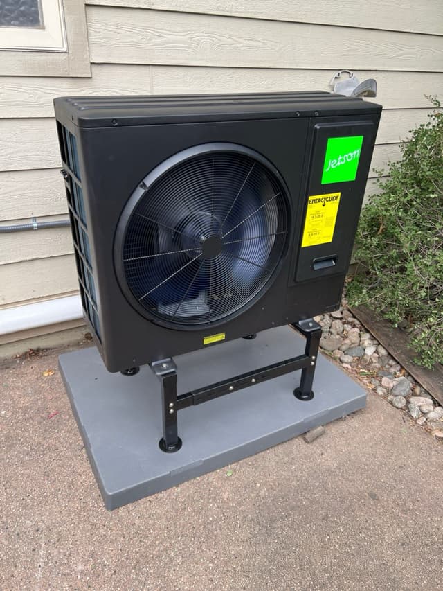 Black Jetson Air 3-ton heat pump condenser on a metal riser stand along the side of a tan vinyl-sided house in Fort Collins, CO.