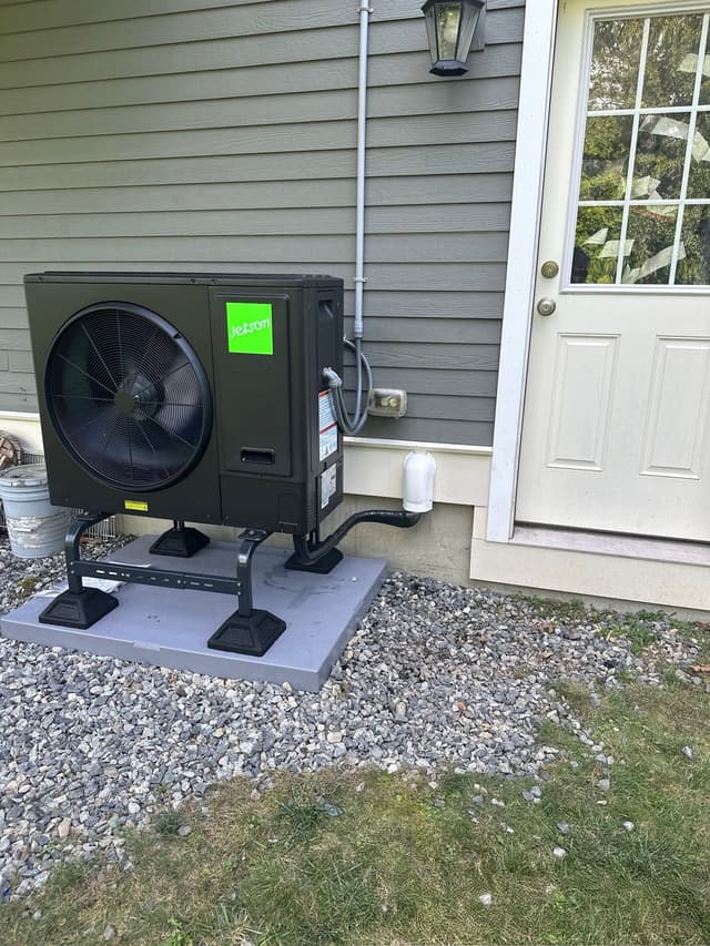 Jetson Air 3-ton outdoor heat pump condenser installed along the gray siding of a Framingham home, with conduit running to a disconnect box and a white entry door visible to the right.