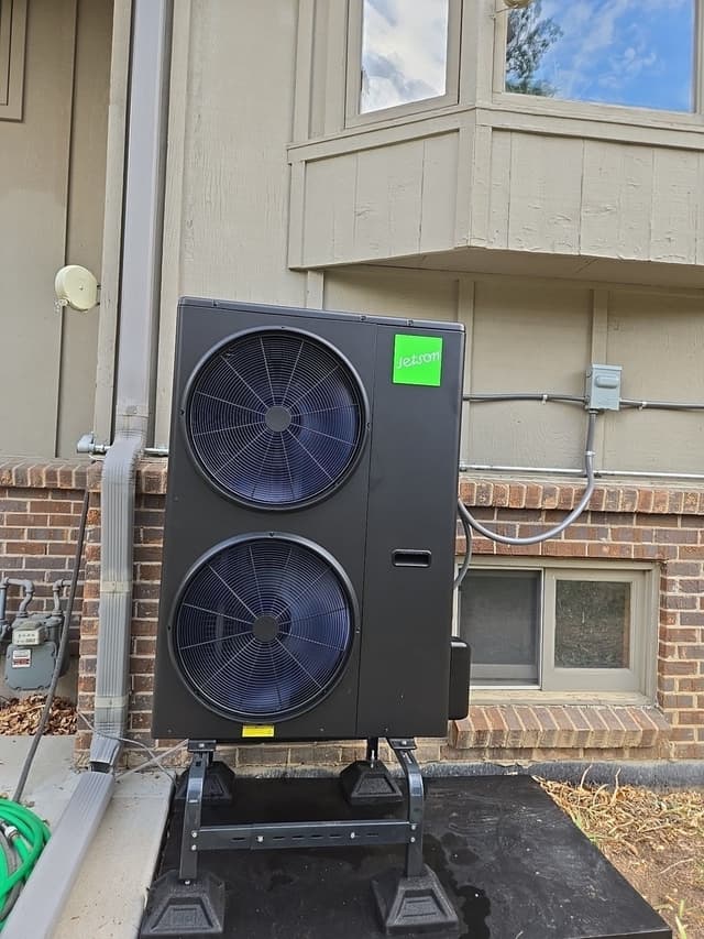 A 4-ton Jetson Air central ducted heat pump installed on an elevated stand outside a multi-story brick-and-siding residential building in Greenwood Village, with conduit and line sets running along the brick wall.