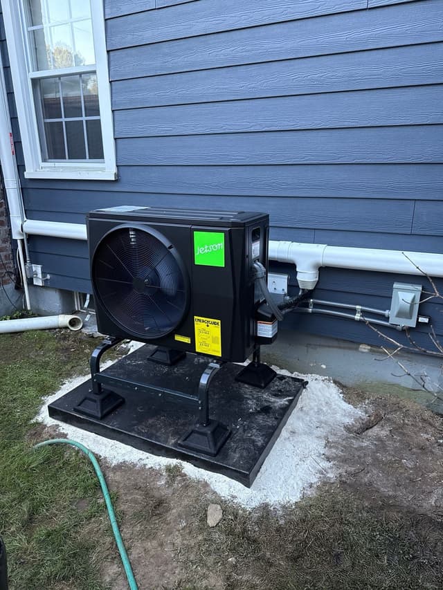Black Jetson Air outdoor heat pump unit installed on the side of a blue-gray lap-sided house in Hartsdale, with refrigerant lines and electrical conduit routed into the wall.