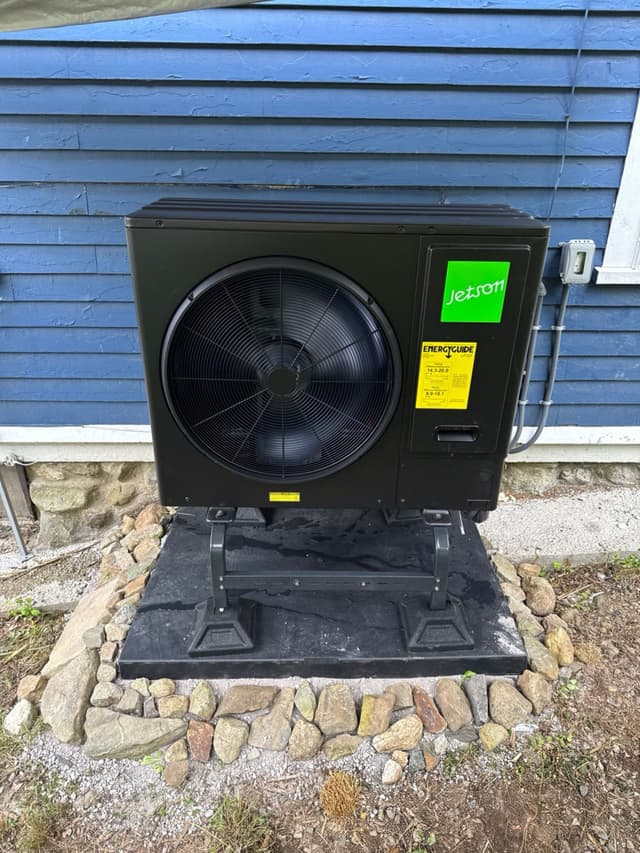 Jetson Air 3-ton outdoor heat pump on a stand and rubber pad, framed by a border of natural fieldstones, mounted against blue vinyl siding in Haverhill, MA.