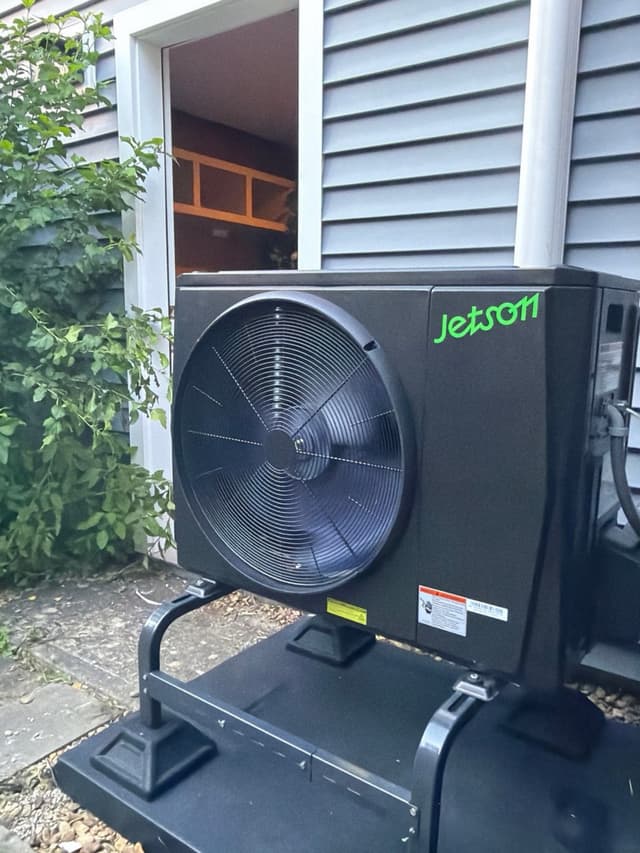 Jetson Air 2-ton central ducted heat pump mounted on a black equipment stand on a gravel pad alongside a gray-sided house in Hopkinton, MA.