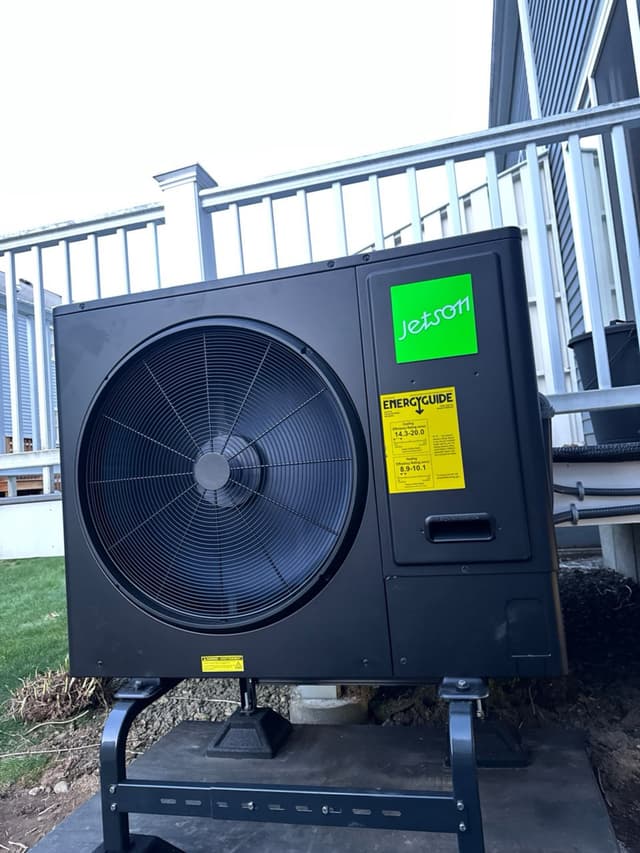 Jetson Air 3-ton central ducted heat pump mounted on a black metal riser on a gravel pad beside a residential deck in Hopkinton, MA.