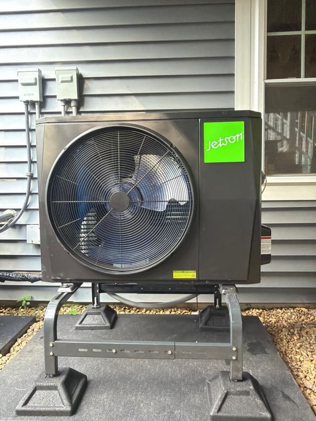 A 2-ton Jetson Air central ducted heat pump mounted on a metal riser stand against gray vinyl siding in Hopkinton, MA, with a disconnect box and conduit neatly installed above.