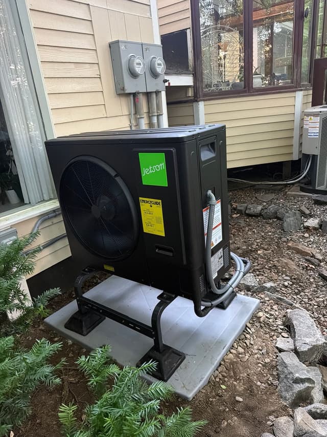 Black Jetson Air 3-ton heat pump condenser installed along the side of a house in Hudson, with refrigerant lines running down the right side and a second condenser unit visible in the background.