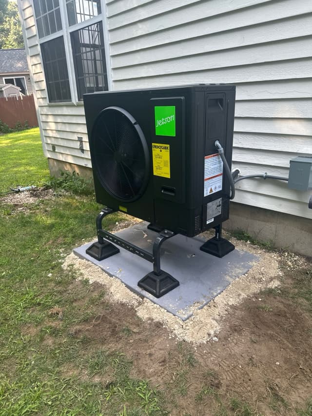 Black Jetson Air 3-ton heat pump outdoor unit mounted on metal riser stands on a concrete pad along the side of a light gray vinyl-sided house in Hudson, MA, with line set conduit running up the wall to a disconnect box.