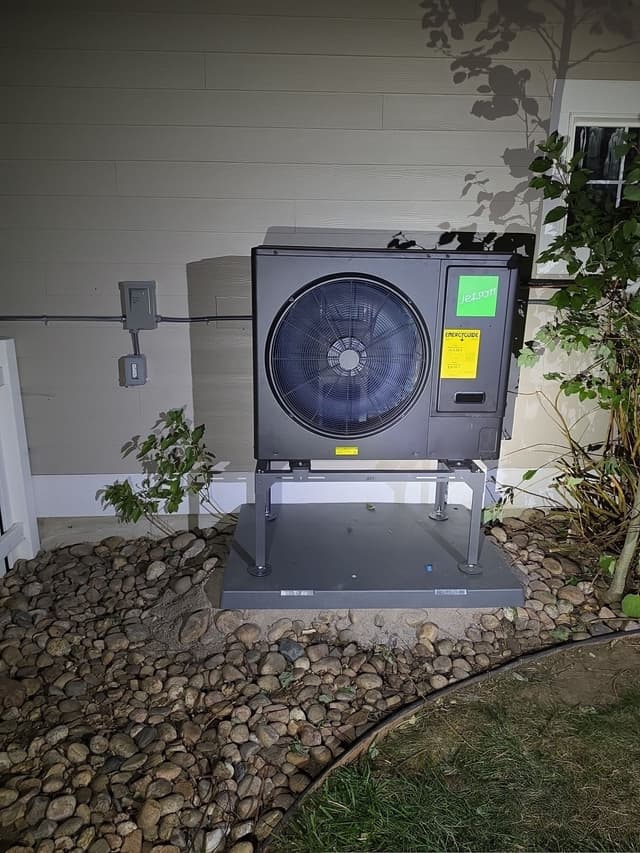 Black Jetson Air 3-ton heat pump installed beside a Lafayette home exterior, with an electrical disconnect box mounted on the siding, photographed at night.