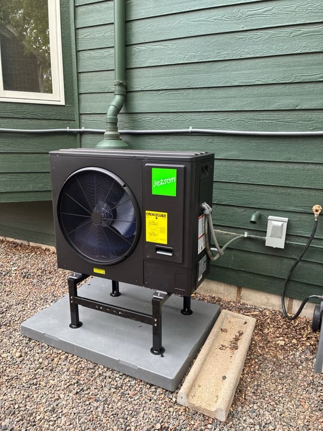 Jetson Air 3-ton heat pump condenser on an elevated metal stand against green lap siding, with electrical conduit and disconnect box mounted on the house wall, Lafayette, CO.