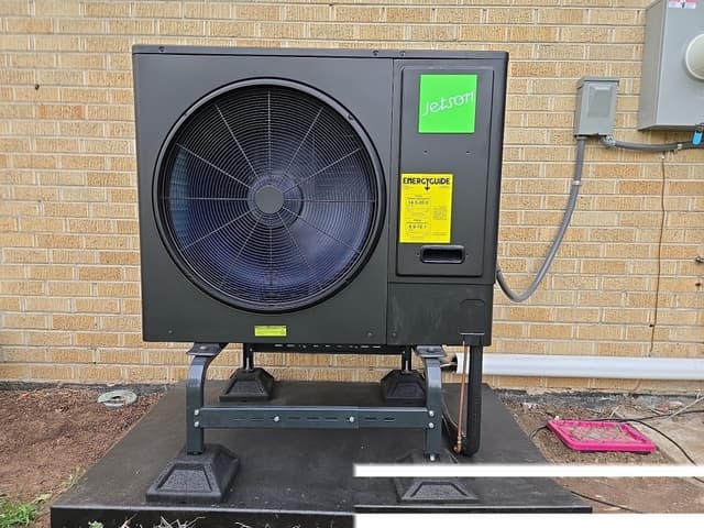Jetson Air 3-ton central ducted heat pump condenser mounted against a tan brick wall at a Lakewood home, with electrical conduit and refrigerant lines running to the wall and a disconnect box above.