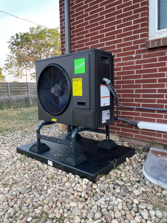 Black Jetson Air 3-ton heat pump condenser mounted on a metal stand over river-rock gravel beside a red brick house in Lakewood, CO, with electrical conduit running neatly into the wall.