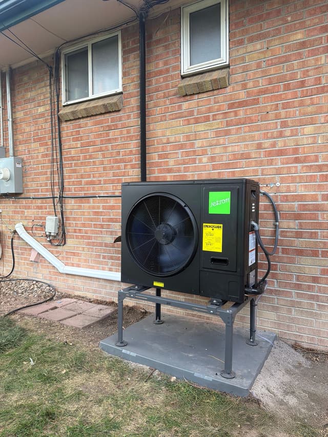 Black Jetson Air 3-ton heat pump condenser mounted on an elevated stand against the brick exterior wall of a home in Lakewood, with refrigerant lines and conduit running up the wall.