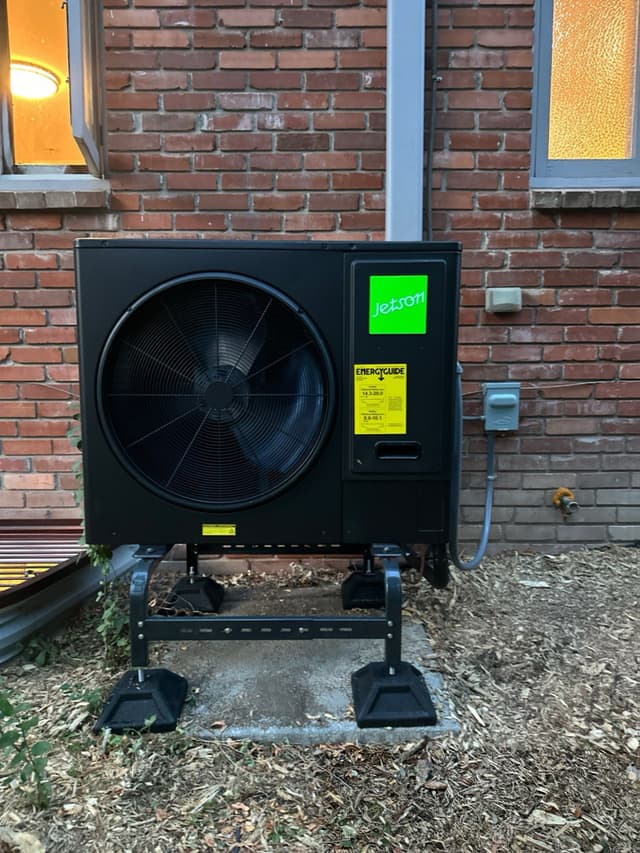 Jetson Air 3-ton central ducted heat pump mounted against a red brick wall between two windows, with electrical conduit running to a disconnect box, Lakewood.