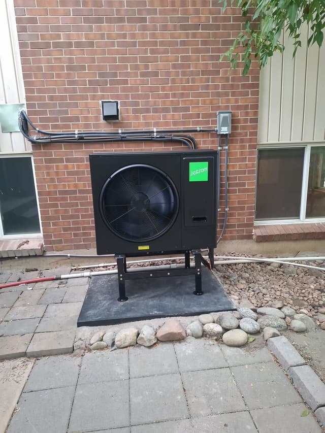 3-ton Jetson Air central ducted heat pump mounted on a stand against a red brick exterior wall on a paver patio in Lakewood, with conduit running neatly to a disconnect box.