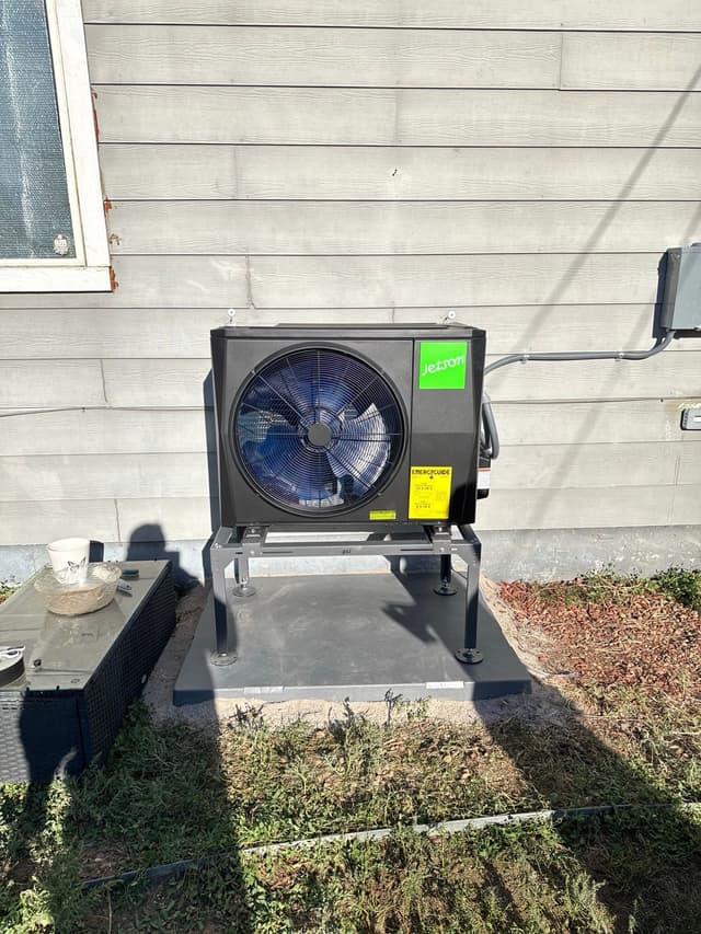Black Jetson Air 2-ton heat pump mounted on a raised metal stand against gray house siding in Lakewood, with electrical conduit running to a disconnect box on the wall.