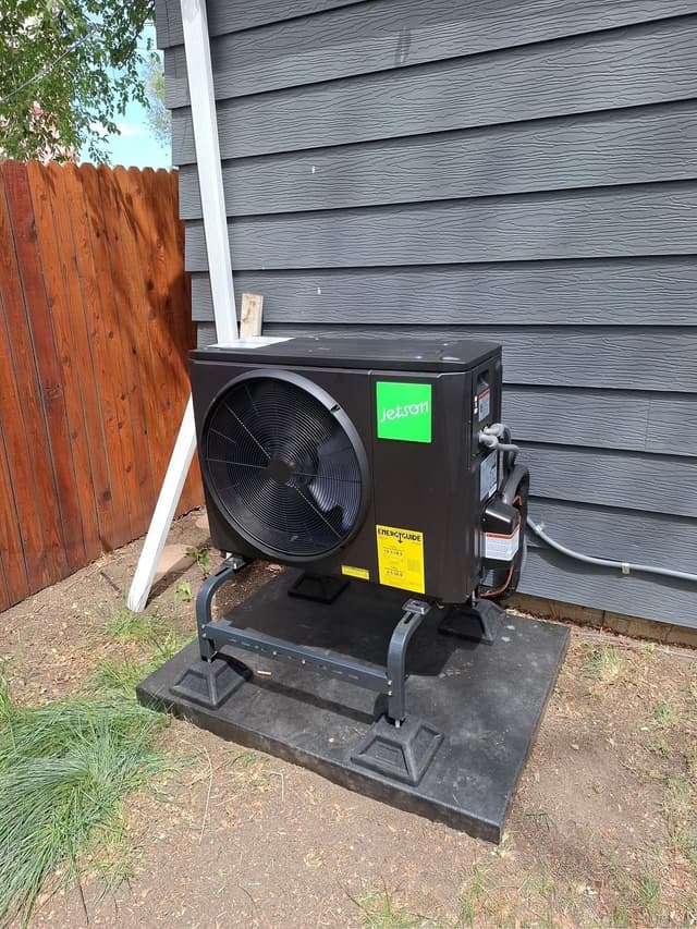 Black Jetson Air 2-ton heat pump condenser on a stand against grey lap siding on a Lakewood, CO home, with refrigerant lines and a wood fence visible to the side.