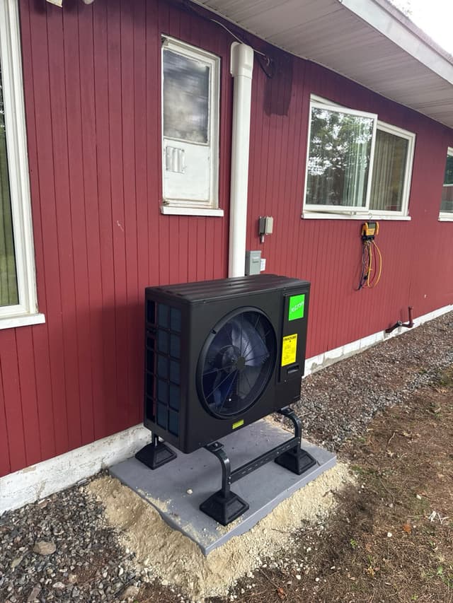 Black Jetson Air 3-ton heat pump condenser on a metal riser stand and composite pad along the red vertical-board-sided exterior wall of a home in Lexington, MA.