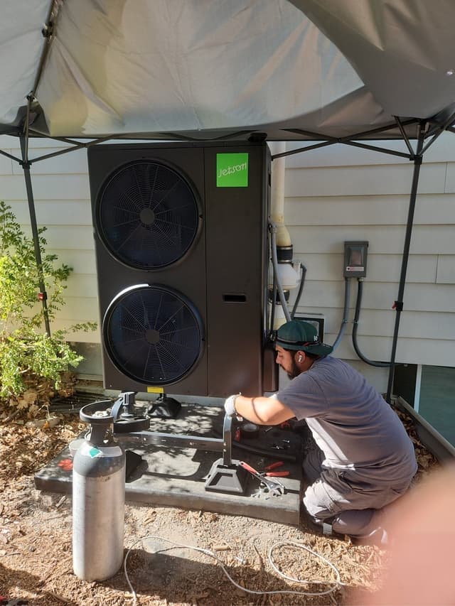 Technician crouched and working on a 4-ton Jetson Air heat pump beside a house in Columbine, Littleton, CO, with a shade canopy overhead and refrigerant equipment on the ground nearby.