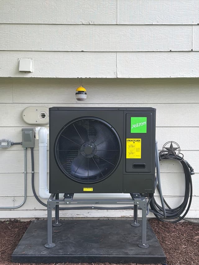 Jetson Air 3-ton central ducted heat pump installed against light grey lap siding on a mulched bed in Littleton, MA, with white line-set cover and disconnect box mounted on the wall.