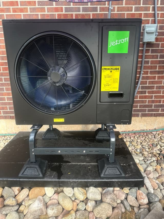 Jetson Air 3-ton central ducted heat pump mounted on a metal stand over a concrete pad with river rock landscaping, brick wall with disconnect box visible to the right, Littleton home.