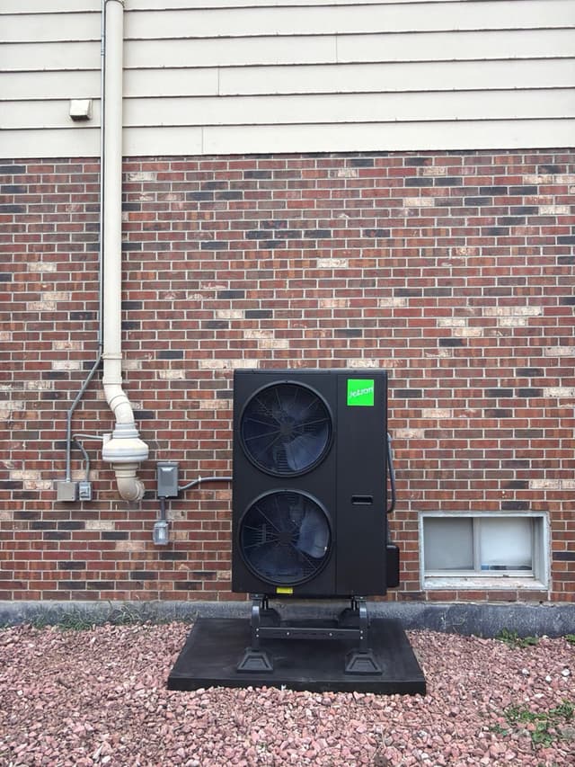 A black Jetson Air 4-ton central ducted heat pump mounted on a gravel bed against a brick exterior wall in Columbine, Littleton, CO, with white PVC pipe and electrical disconnect box visible.
