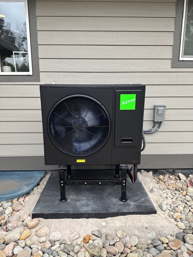 Black Jetson Air 3-ton central ducted heat pump condenser installed along the side of a gray-sided house in Littleton, MA, with disconnect box and conduit run on the wall beside it.