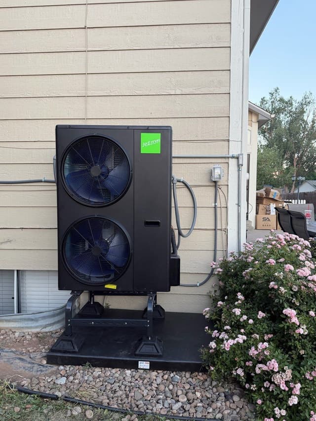 Jetson Air dual-fan heat pump condenser on a metal stand against a tan lap-siding home in Littleton, Colorado, with electrical conduit and refrigerant lines running neatly to a disconnect box.