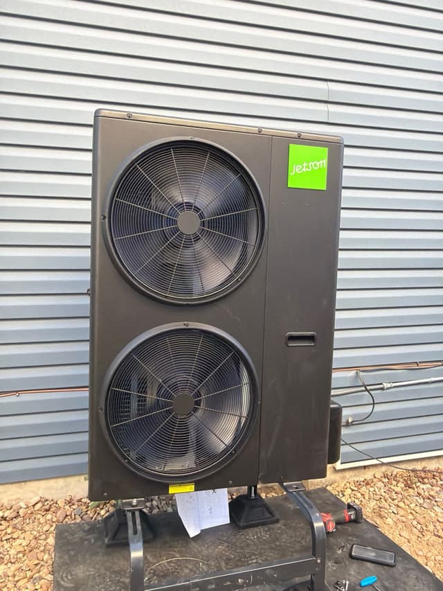 Black Jetson Air 4-ton heat pump with dual stacked fans installed against a grey corrugated metal wall in Littleton, CO, with refrigerant lines and electrical conduit running along the wall.