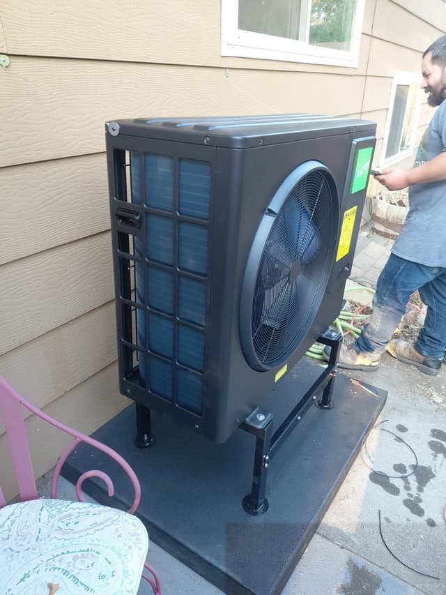 A black Jetson Air heat pump unit installed against tan house siding in Longmont, with a smiling bearded technician partially visible to the right.