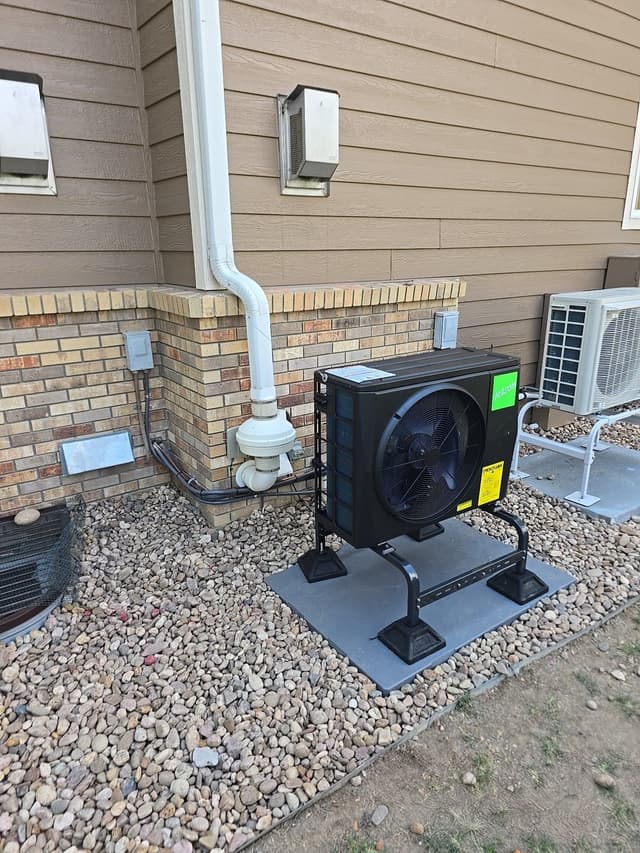 Black Jetson Air 2-ton central ducted heat pump condenser installed against the side of a tan lap-siding home with brick foundation, surrounded by decorative rock landscaping in Longmont, Colorado.