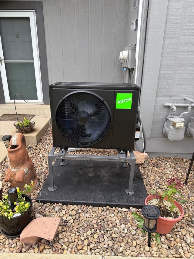 Black Jetson Air heat pump condenser installed in a river-rock landscape bed alongside a house in Longmont, Colorado.