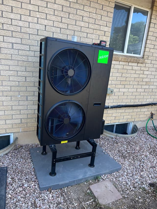 Black Jetson Air 4-ton central ducted heat pump mounted against a tan brick exterior wall on a concrete pad with gravel landscaping, Longmont, CO.