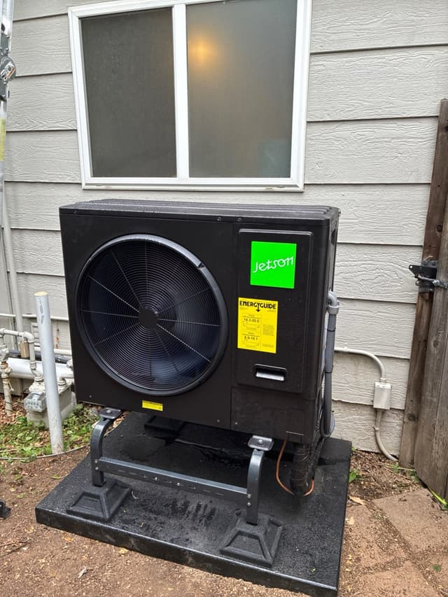 Jetson Air 3-ton outdoor heat pump unit mounted on a black pad with riser legs along the side of a gray house in Longmont, Colorado, with refrigerant lines and conduit running down the side panel.