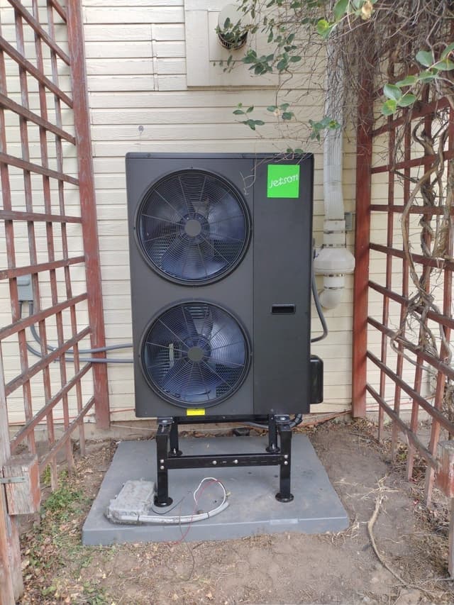 Black Jetson Air 5-ton central ducted heat pump installed against beige lap siding in Loveland, CO, flanked by rustic wooden trellis panels with vines.