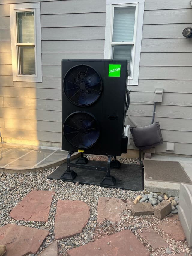A large black Jetson Air dual-fan 4-ton heat pump condenser on a gravel pad against gray lap siding, with line set and electrical conduit running up to a wall-mounted disconnect box, at a home in Loveland, CO.