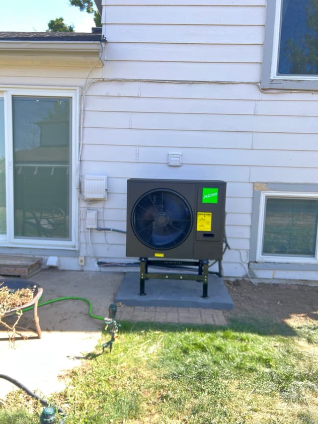 Black Jetson Air 3-ton central ducted heat pump condenser mounted against white vinyl siding of a home in Loveland, CO, with line sets running along the exterior wall.