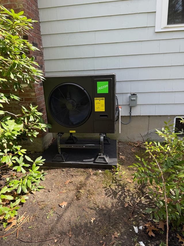 Black Jetson Air 3-ton outdoor heat pump unit installed at ground level against a brick foundation and white vinyl siding on a Medfield, MA home, flanked by rhododendron bushes.