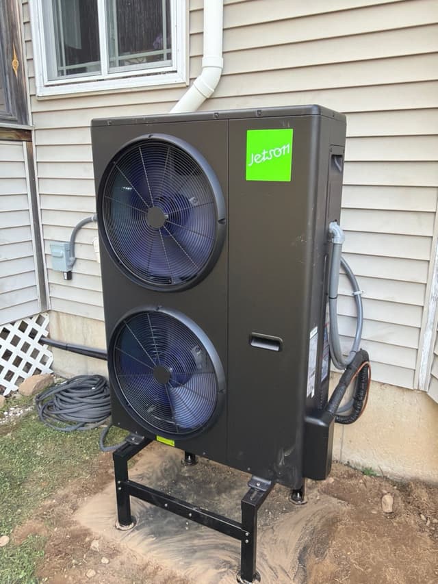 Jetson Air 4-ton heat pump on a black metal stand beside a house with tan vinyl siding in Medway, MA, with conduit and refrigerant lines running along the right side and an electrical disconnect box mounted on the wall.
