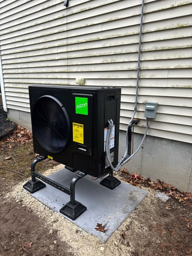 Jetson Air 3-ton heat pump mounted on a metal stand over a concrete pad against vinyl siding of a home in Norfolk, MA, with conduit and refrigerant lines running up to a disconnect box.