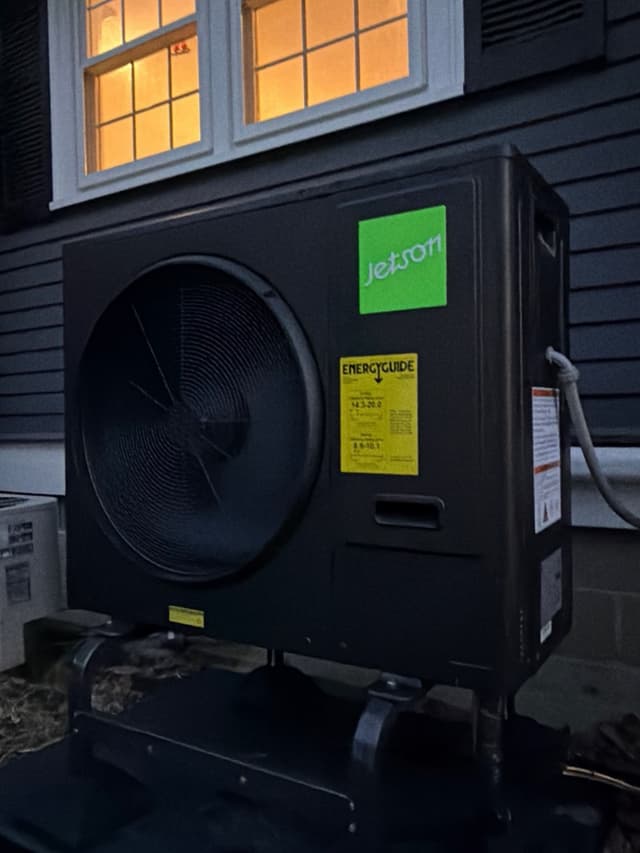 Black Jetson Air 3-ton heat pump on a low equipment stand against dark gray horizontal siding, with a warm lit window above and a white refrigerant line exiting the right side, photographed at dusk.