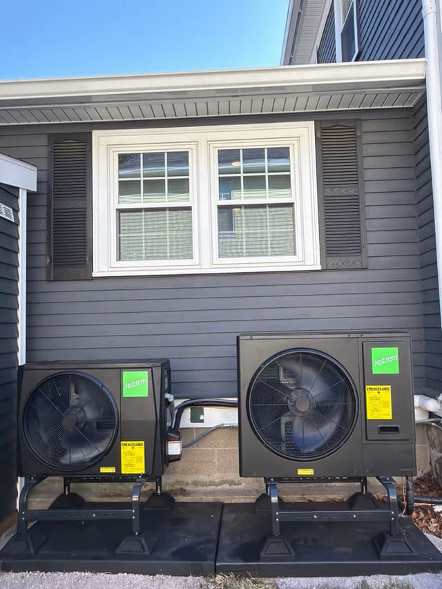 Two black Jetson Air outdoor heat pump condensers mounted side by side on black metal stands along the side of a gray-sided home in North Andover, MA.