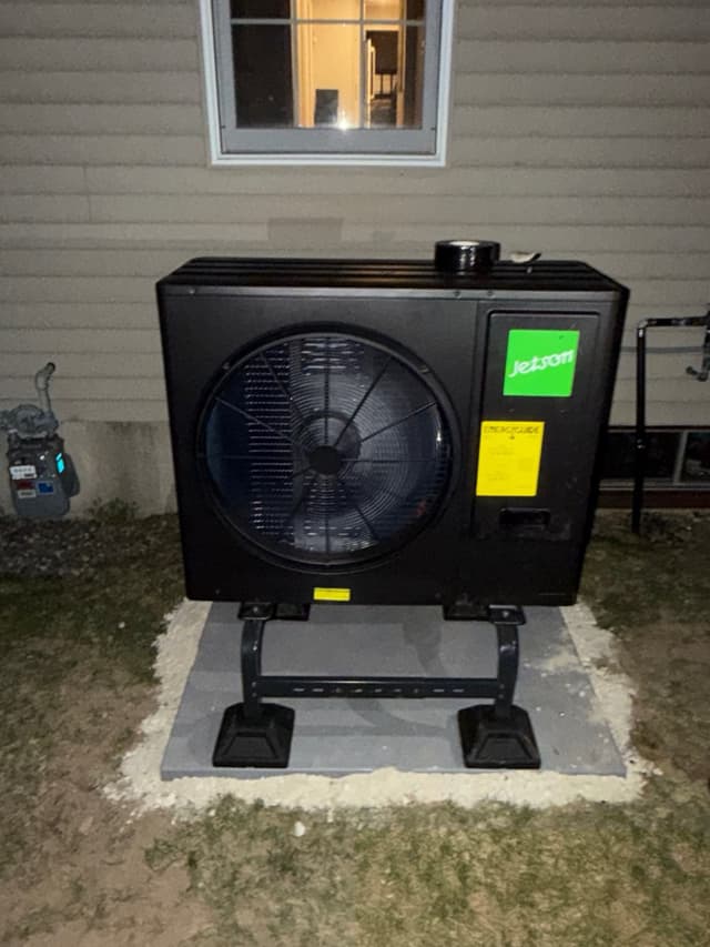 Jetson Air 3-ton central ducted heat pump outdoor unit mounted on a metal stand over a gravel pad against gray vinyl siding of a North Andover, MA home, photographed at night.