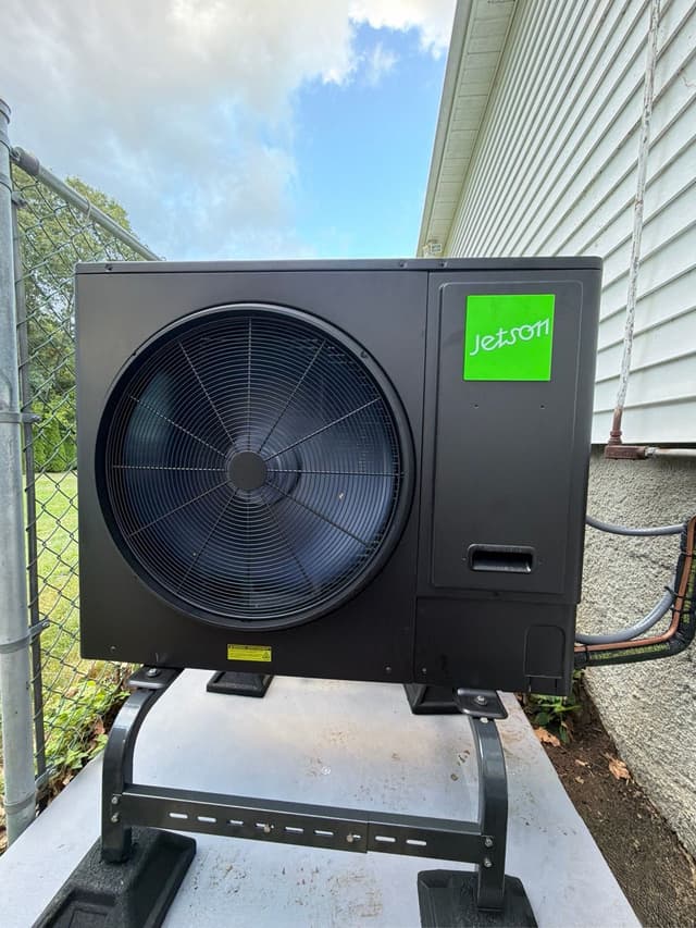 A black Jetson Air 3-ton central ducted heat pump installed on the side of a light-gray vinyl-sided house in North Attleborough, MA, with a chain-link fence and lawn visible to the left.