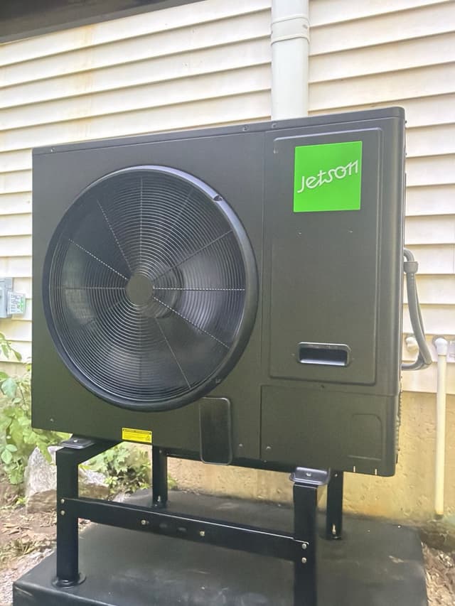 Jetson Air 3-ton heat pump on an elevated stand against the exterior wall of a Northbridge, MA home, fully installed with refrigerant lines and PVC pipe visible on the wall.