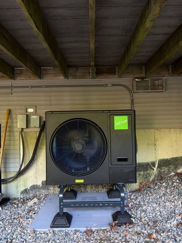 3-ton Jetson Air central ducted heat pump installed beneath a low deck with exposed wooden joists, conduit running up the house wall, and an electrical disconnect box mounted on the siding in Norton, MA.