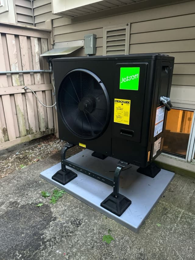 Jetson Air 3-ton heat pump on a composite pad with anti-vibration standoffs in a narrow side yard between a wooden fence and house siding, Oxford, MA.