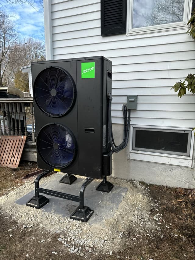 A large black dual-fan heat pump condenser on a metal equipment stand and concrete pad beside a white-sided house in Peabody, MA, with conduit running neatly to a disconnect box on the wall.