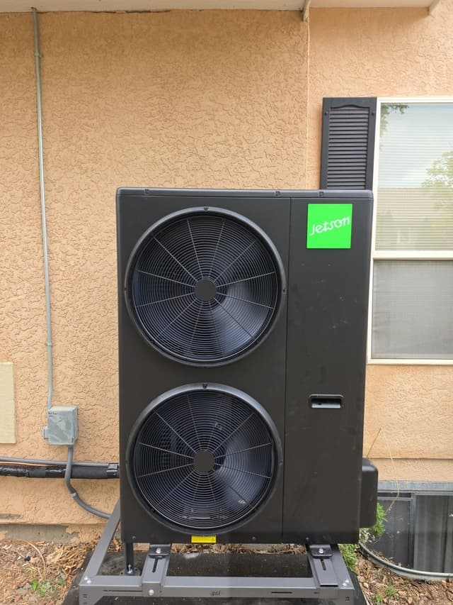 Black Jetson Air 4-ton heat pump condenser mounted against the stucco exterior of a home in Pueblo, Colorado, with conduit running up the wall and lineset exiting from the bottom.