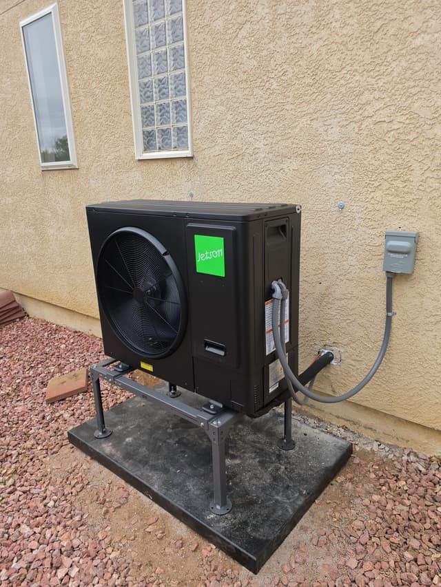 Black Jetson Air 3-ton central ducted heat pump installed against a stucco wall with a glass-block window, red gravel ground, in Pueblo, Colorado.